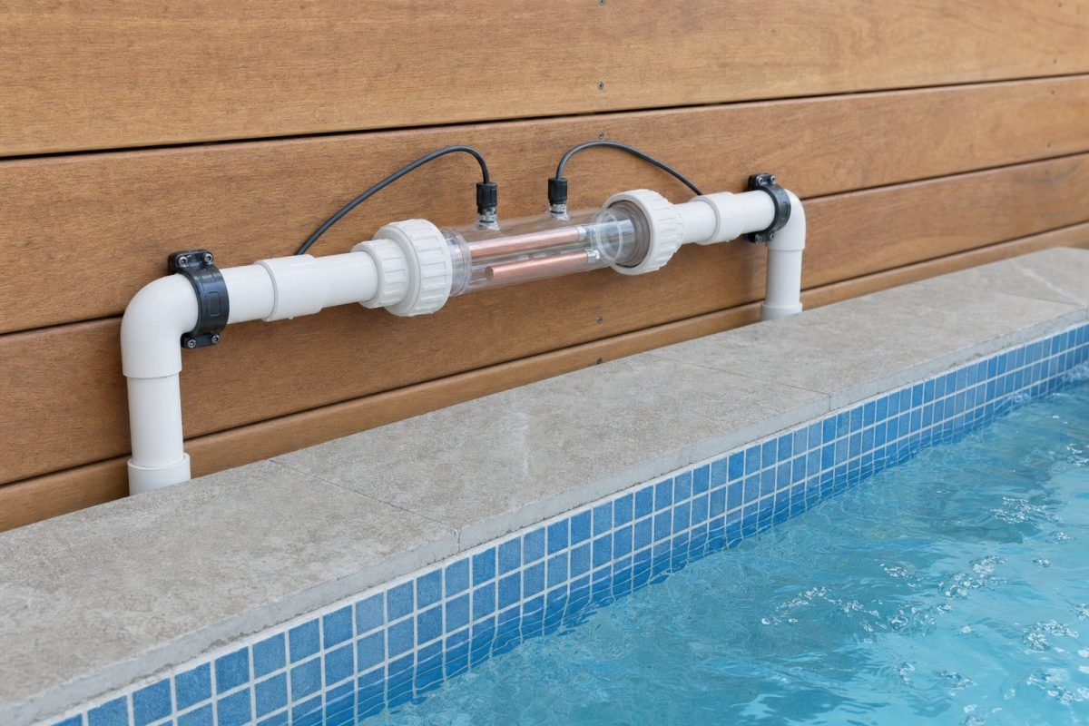 Inline copper-silver ioniser chamber with copper-coloured electrodes fitted to white pipework beside a hot tub