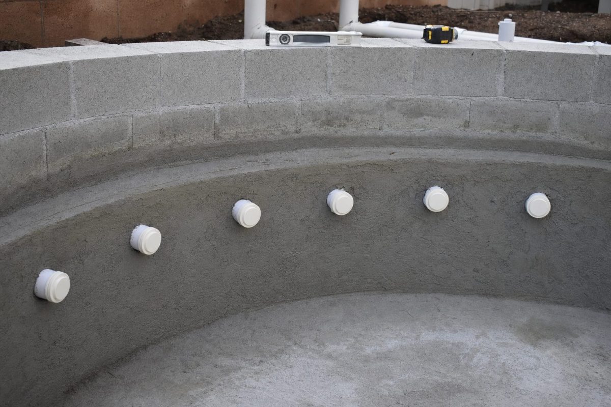 Row of white gunite jet bodies set through the concrete block wall of a DIY hot tub with water and air pipes connected behind
