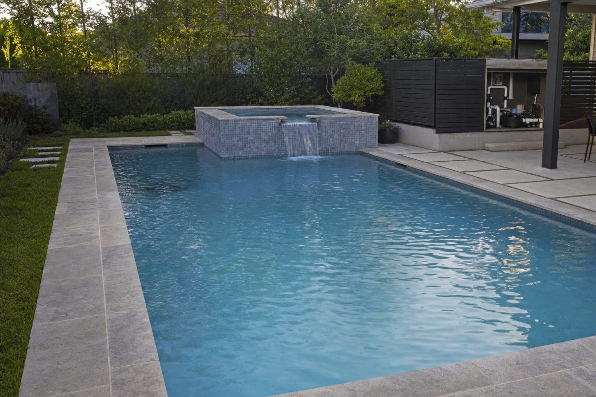 In-ground swimming pool with a raised spillover hot tub at one end and separate equipment behind