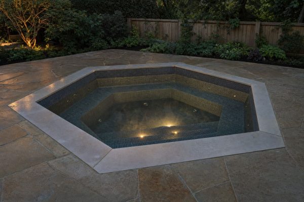 Hexagon hot tub sunk flush into a stone patio at dusk, with dark mosaic tiles, bench seats and a central footwell